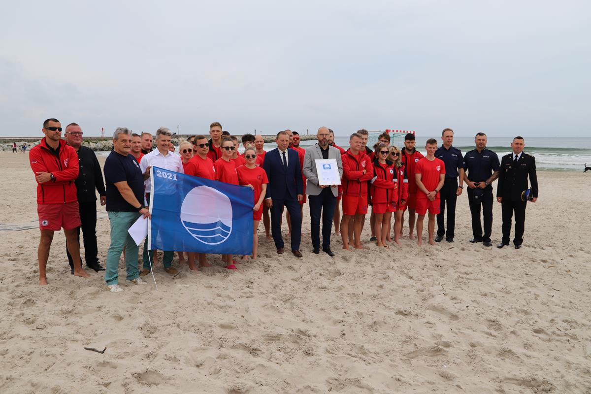 Ratownicy stoją, w tle morze, z przodu piasek, trzymają flagę