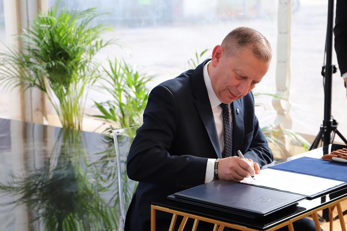 Burmistrz przy stoliku podpisuje dokumenty, w tle kwiat doniczkowy