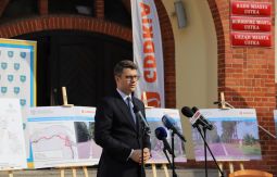 Minister Piotr Muller przemawia, w tle plansze, wejście do ratusza