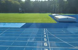 niebieska bieżnia na stadionie lekkoatletycznym, w tle las, kawałek boiska z trawą